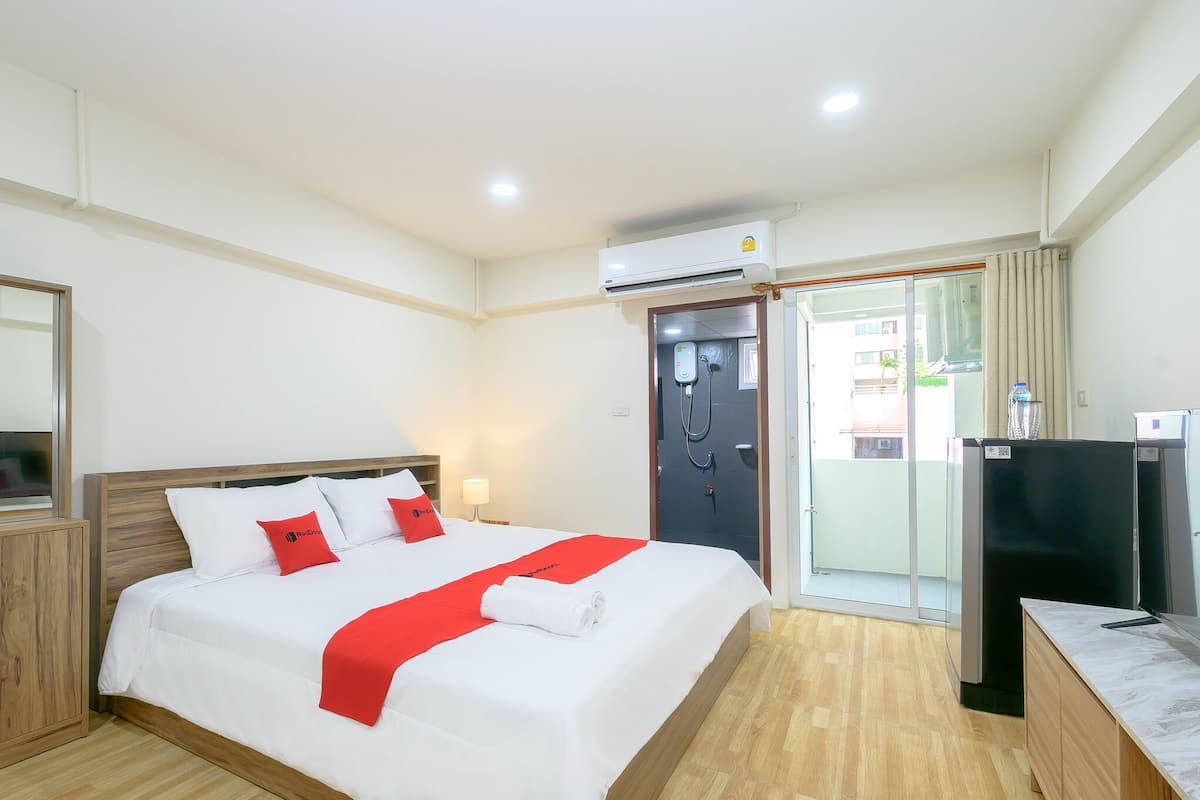 A hotel room with a bed, a desk, an air conditioner, a door, and a view of the outside.