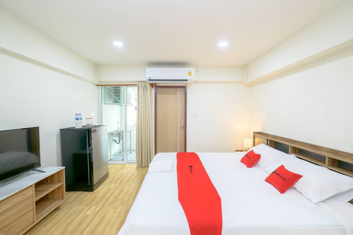 A hotel room with a bed, a TV, a mini-fridge, and an air conditioning unit.