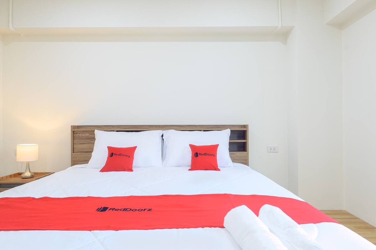 A hotel room with a bed, white bedding, two red pillows, a red blanket, and a nightstand with a lamp.