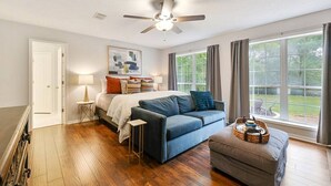 A bedroom with a bed, a blue sofa, a grey ottoman, a ceiling fan, and a large window with curtains.
