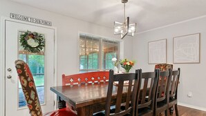 A dining area with a wooden table, six chairs, a red bench, a chandelier, and a door with a wreath.