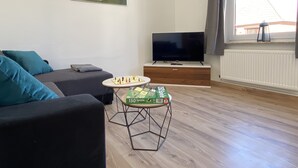 Ett vardagsrum med en soffa, ett litet soffbord med ett brädspel, en tv på ett tv-bord och ett fönster med utsikt över en byggnad.