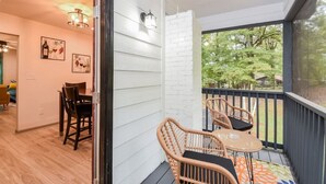 Un balcon avec des fauteuils et une table en osier, des murs recouverts d’un revêtement blanc, et une vue sur la verdure.