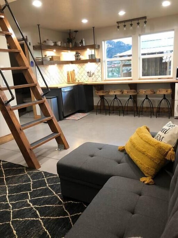 A modern kitchen with a wooden ladder, a grey sofa with a yellow throw pillow, and a view of mountains through the window.