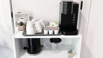 Una estantería blanca con una cafetera, tazas, una tetera, una planta pequeña y un libro.