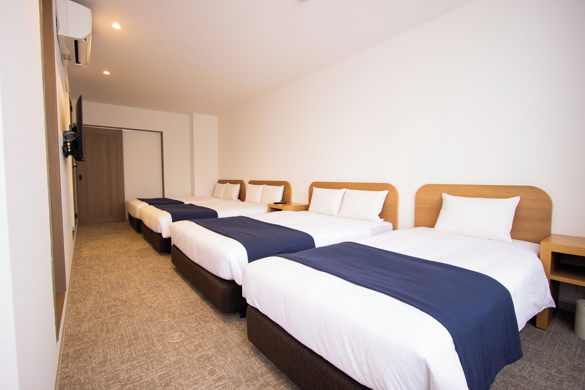 Habitación de hotel con cuatro camas individuales, cada una con cabecero de madera y ropa de cama blanca. El suelo está alfombrado y hay una mesita de noche de madera pequeña con una lámpara en cada cama.