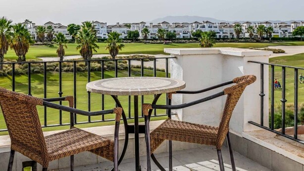 A balcony with wicker furniture, a round table, and a view of a golf course and buildings in the distance.