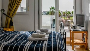 A hotel room with a bed, a TV on a wooden stand, and a balcony with a view of palm trees and buildings.