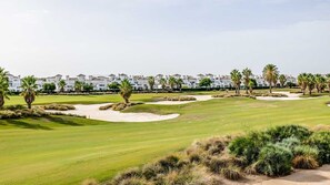 Un parcours de golf avec des bunkers de sable, des palmiers et des bâtiments blancs en arrière-plan.