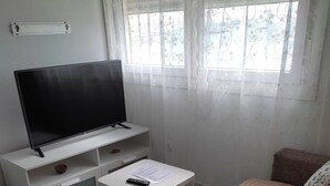 A living room with a flat-screen TV on a white stand, a small white table with a remote and papers, a beige sofa with patterned cushions, and a window with sheer curtains. - Loft con Espectaculares Vistas al Mar, Gibraltar, Marruecos y Bahía de Algeciras (La Línea de la Concepción)