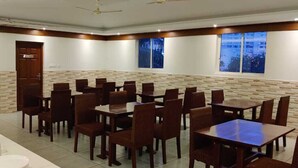 A dining area with multiple tables and chairs, a buffet setup on the left, and a stone wall with a window on the right. - Hotel Burooj (Kanayannur)