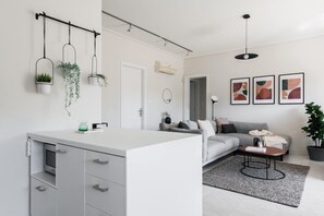 A modern kitchen with a white countertop, grey cabinets, and hanging plants. There is a grey sofa with a coffee table in the living area, and three abstract paintings on the wall. - Magnificent 2BR Apartment in Marousi (Marousi)