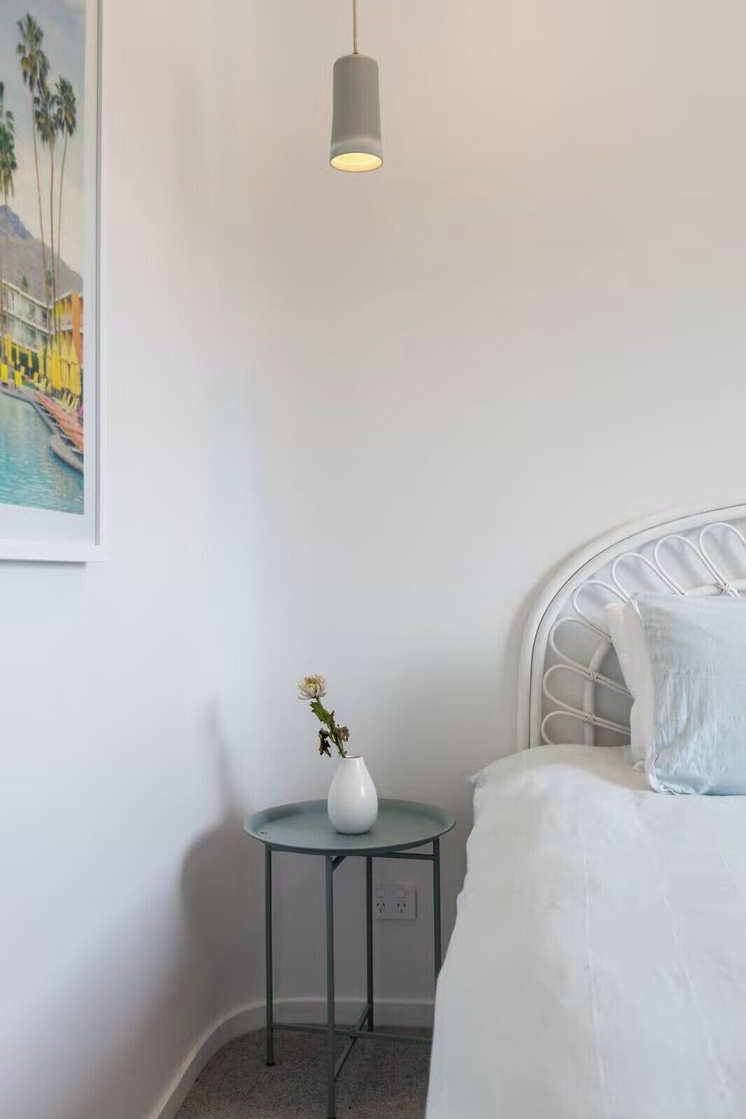 Une chambre à coucher avec un lit, une petite table de chevet sur laquelle se trouve un vase, un tableau au mur et une suspension.