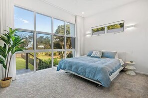 Un dormitorio con una ventana grande, una cama, una planta en maceta y una mesita.