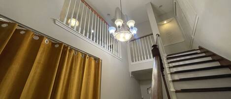 Eine Treppe mit hölzernem Handlauf und weißen Spindeln, die zu einer Empfangshalle im zweiten Stock mit einem Chandelier führt.