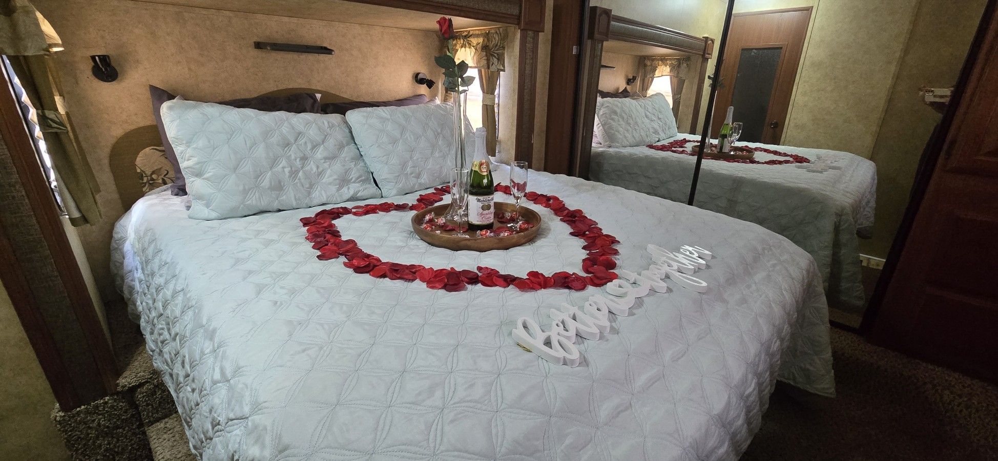 Een tweepersoonsbed met een hartvormige bloemopstelling van rode rozenblaadjes en een fles champagne.