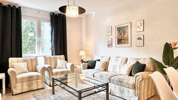Un salon comprenant un canapé, deux fauteuils, une table basse en verre et une lampe. Des tableaux encadrés ornent le mur, et une fenêtre est agrémentée de rideaux.