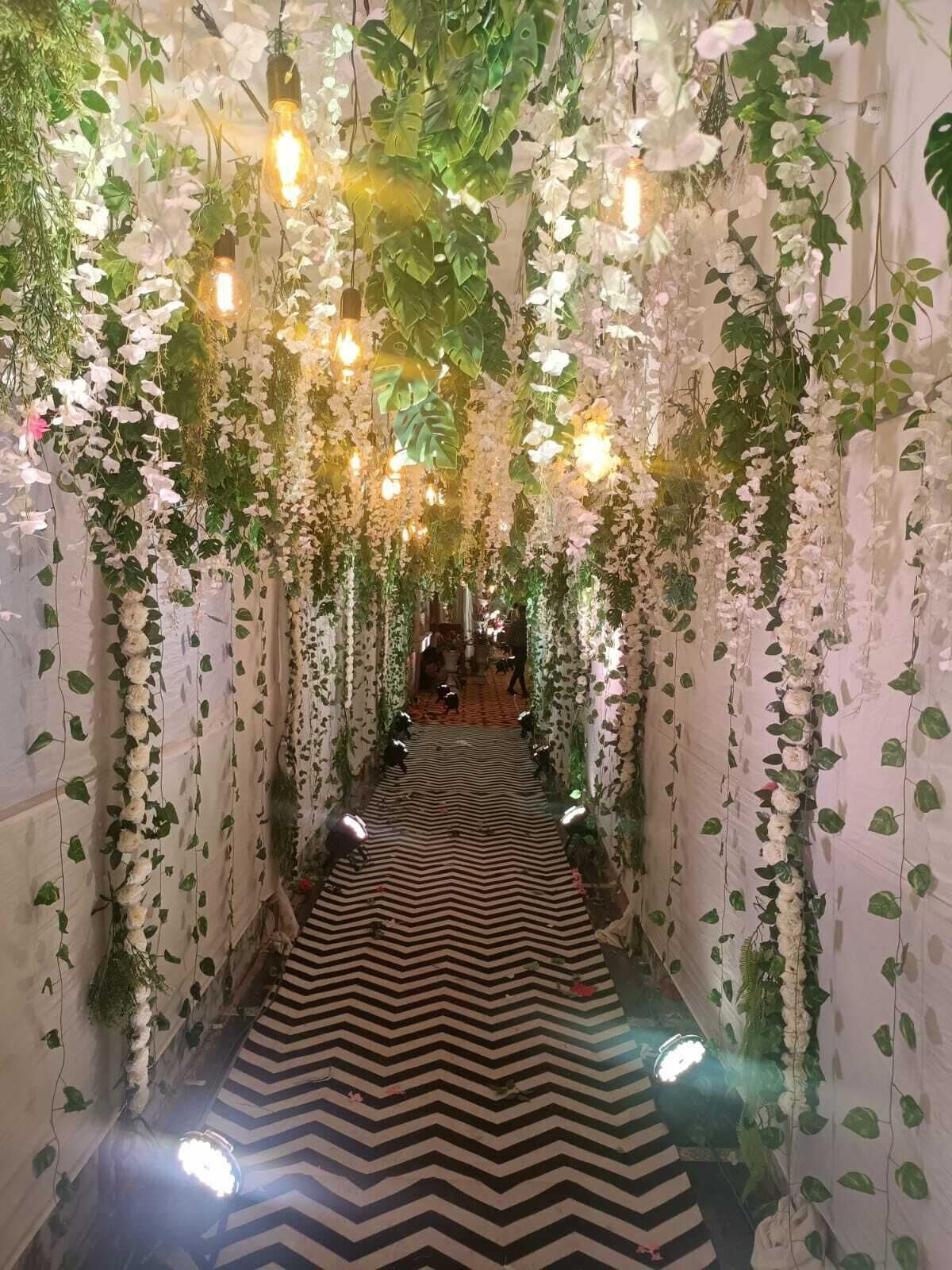 A hallway with a black and white zigzag patterned floor, surrounded by walls covered in green foliage and white flowers, with hanging light bulbs.