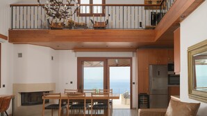 Un vaste espace de vie comprenant une table à manger en bois, un canapé et une vue sur la mer grâce à de grandes portes coulissantes.