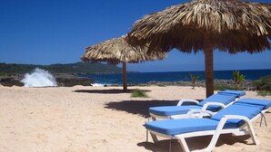 Ein Strand mit Reetdachschirmen, Liegestühlen und einer felsigen Küstenlinie.