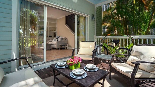 A covered patio with wicker furniture, a dining table set with plates and a flower vase, and a bicycle. - Doc`s Paradise (Key West)