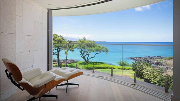 Een moderne kamer met een grote glazen deur die leidt naar een balkon met uitzicht op de oceaan en een boom.