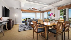 A modern living room with a dining area, large windows, a ceiling fan, and a view of the ocean. - Wai'ula'ula C102 Three Bedroom (Kamuela)
