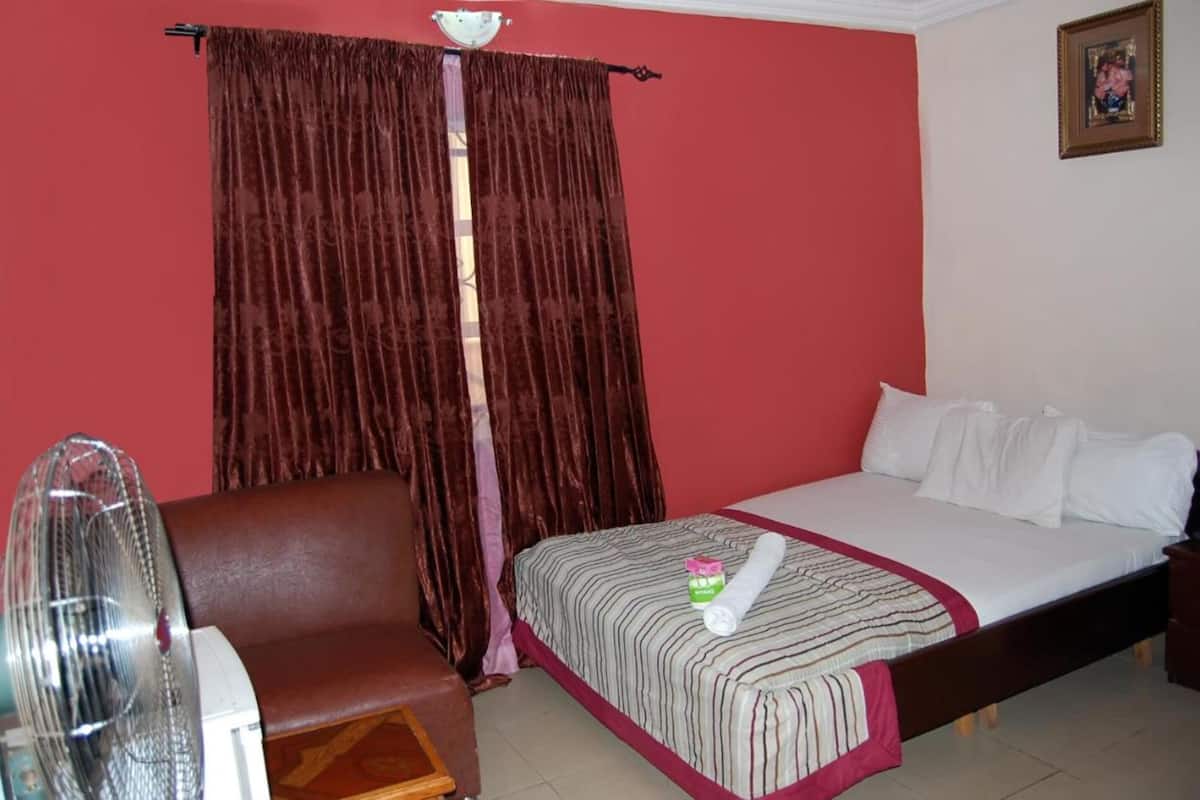 A hotel room with a bed, a chair, a fan, and a curtain.