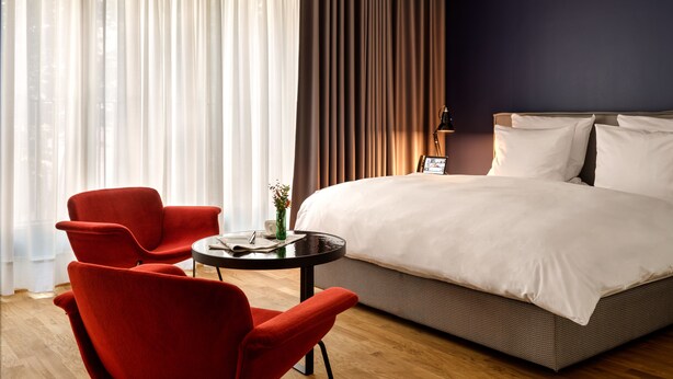 Habitación de hotel con una cama grande, una mesita redonda y una silla, y vistas al exterior a través de cortinas de tela ligera.