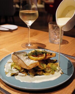 A plate of food with a fish fillet, lemon slices, and potatoes, accompanied by a glass of white wine. - Boston Apartments HH (Hamburg)