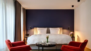 Una habitación de hotel moderna con una cama grande, dos sillas rojas, una mesita redonda y una mesita de noche con lámparas.
