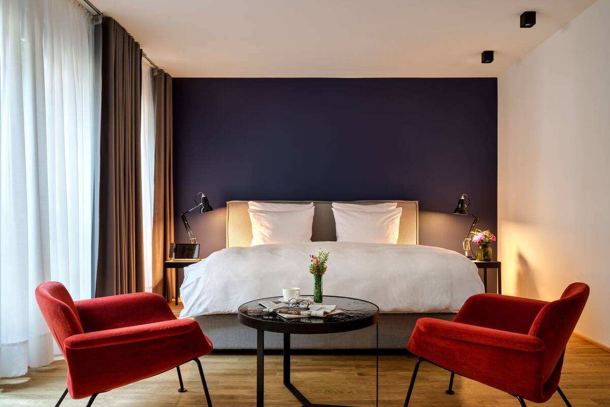 A modern hotel room with a large bed, two red chairs, a small round table, and a nightstand with lamps.