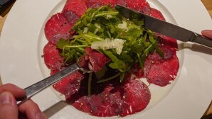 Un plato de carne cruda con vegetales verdes, un bol de verduras y una copa de vino sobre una mesa de madera.