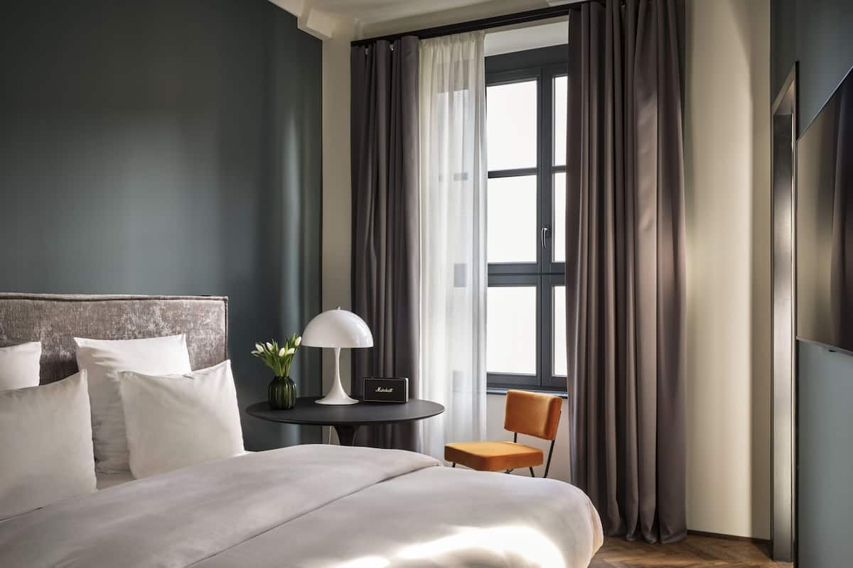 A modern hotel room with a large bed, a small table with a lamp, a chair, and a window with curtains.