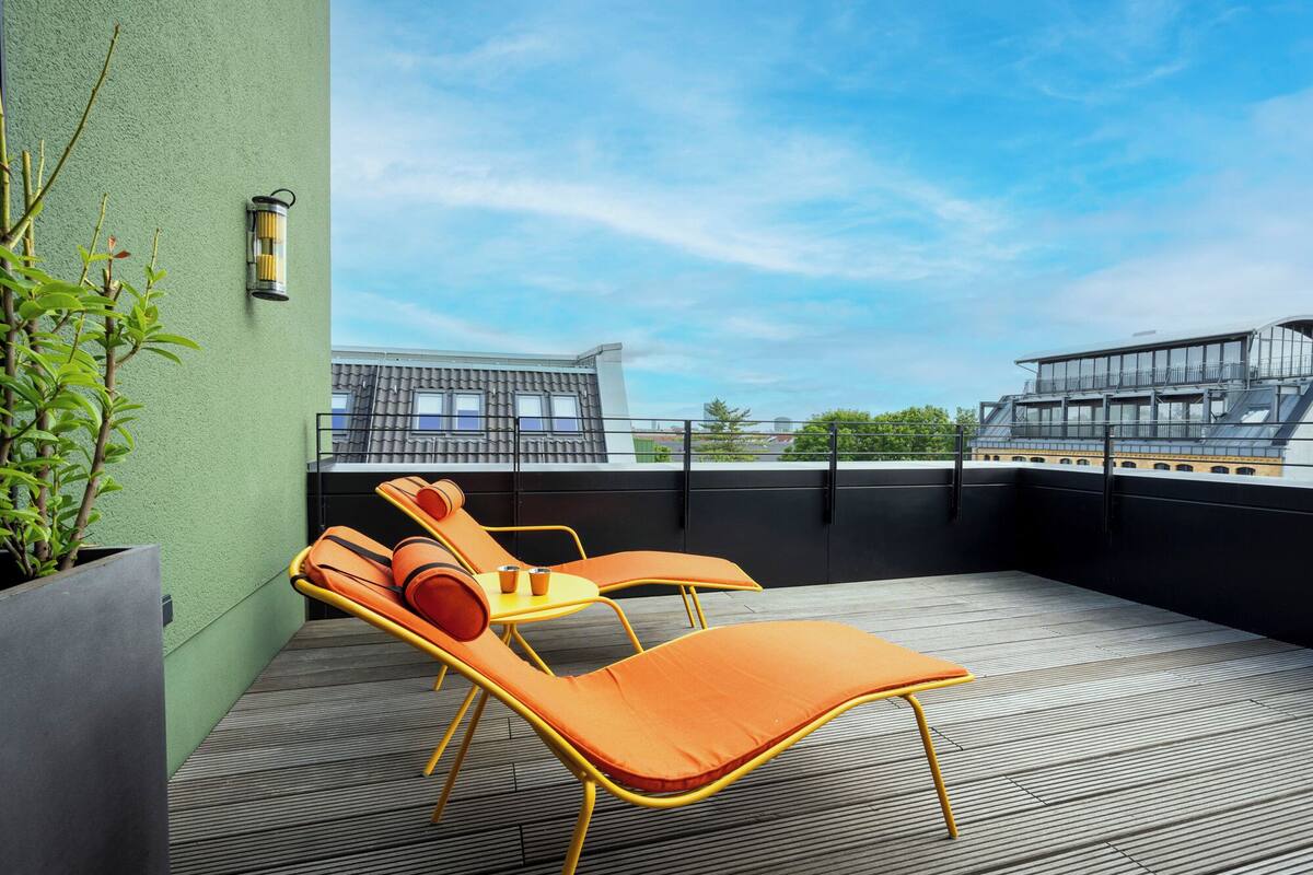 A rooftop terrace with two orange lounge chairs, a potted plant, and a wall-mounted light fixture.