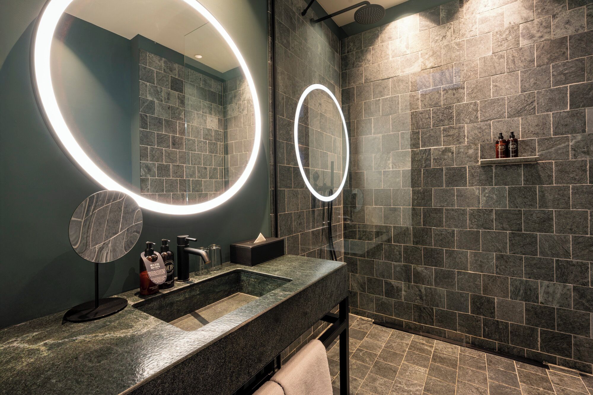 Un baño moderno con encimera de granito, dos espejos iluminados, zona de ducha con pared de azulejos y un estante con artículos de aseo.