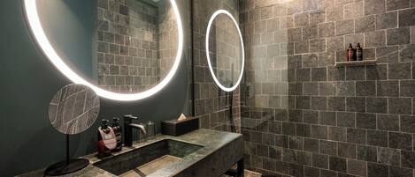 Un baño moderno con encimera de granito, dos espejos iluminados, zona de ducha con pared de azulejos y un estante con artículos de aseo.