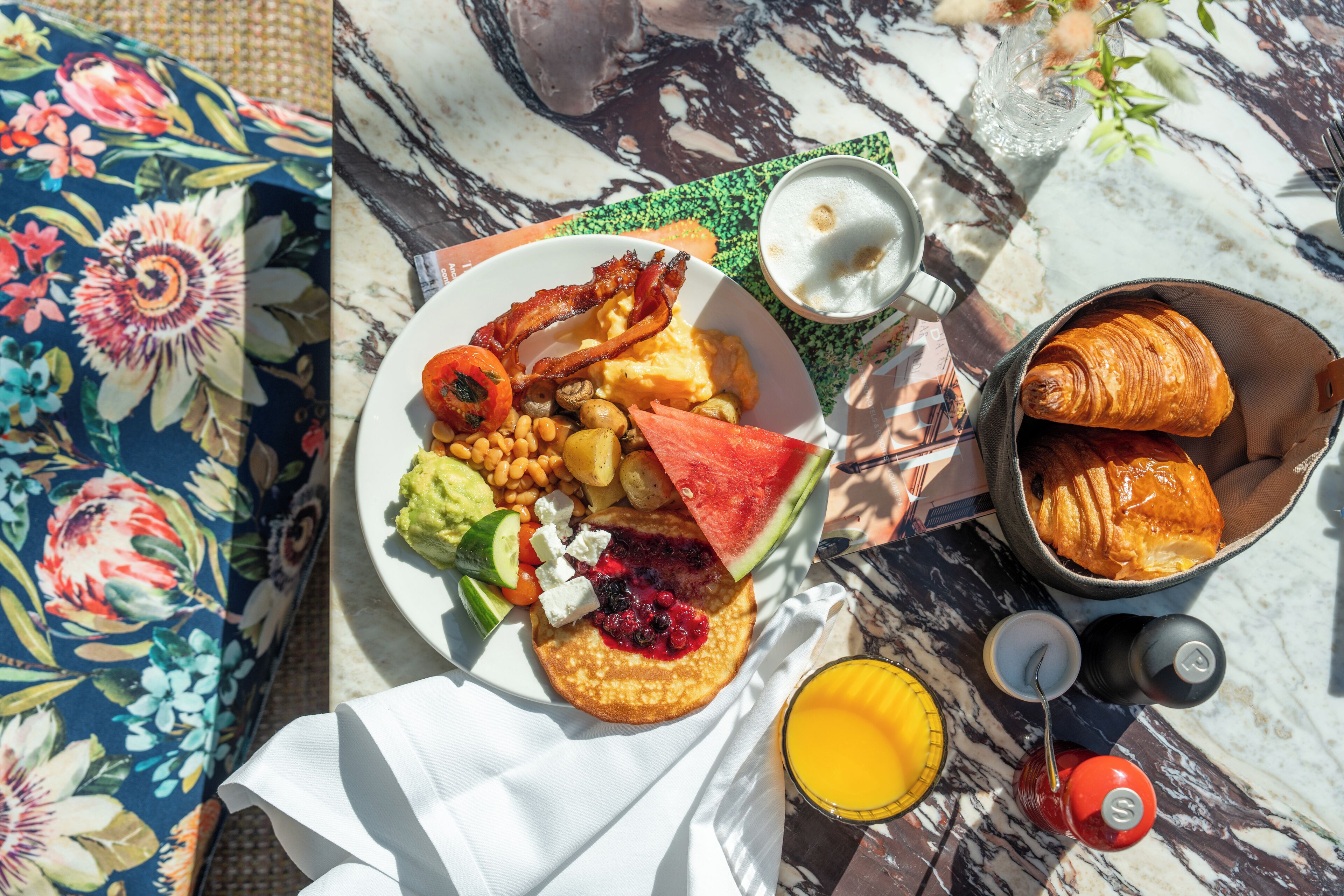A breakfast spread on a table with a floral tablecloth, including pancakes, bacon, eggs, toast, fruit, and a croissant basket.