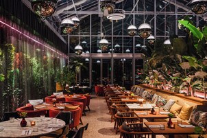 Un ristorante con tavoli rotondi e sedie rosse, un soffitto alto con lampade a sospensione e una parete ricca di piante.