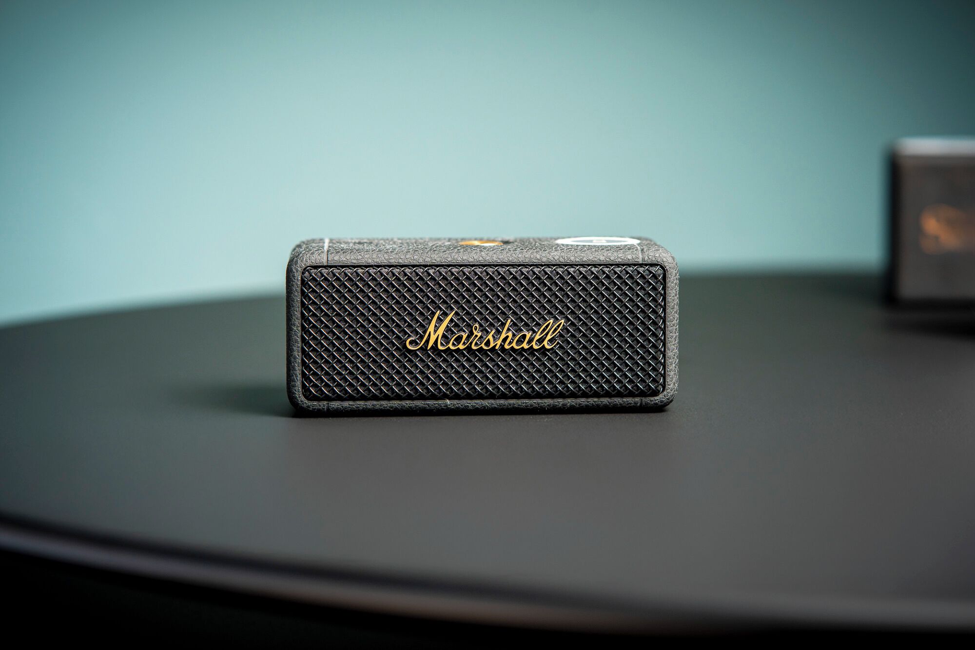 Un altavoz pequeño y texturizado con el nombre de la marca "Marshall", sobre una superficie oscura frente a un fondo verde azulado.
