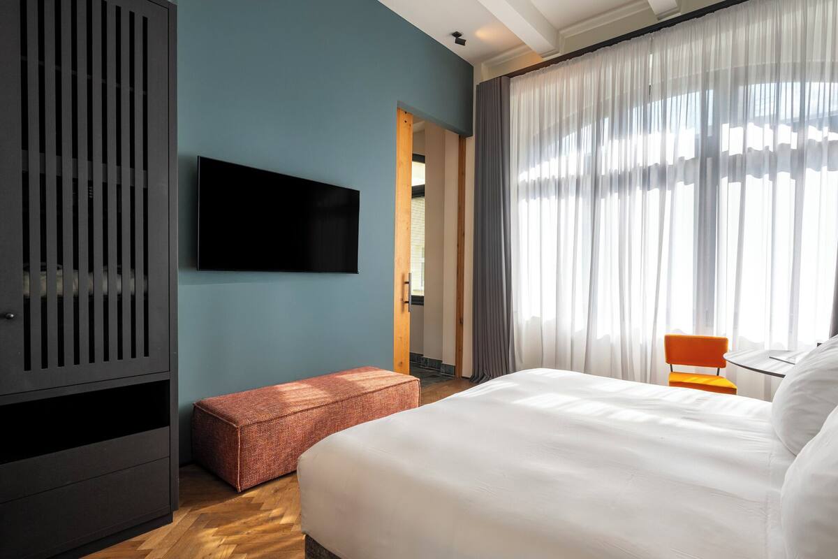 A hotel room with a large bed, a television mounted on the wall, a wooden bench, and a window with sheer curtains.