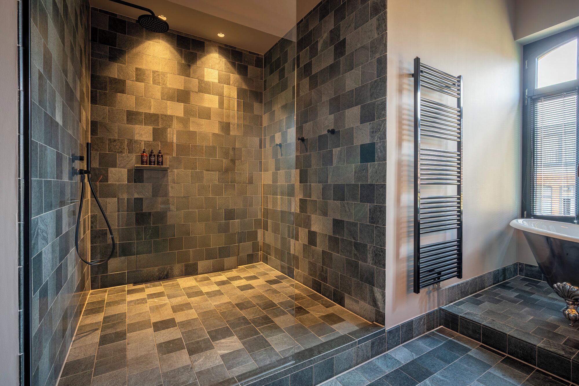 Un baño moderno con suelo y paredes de azulejos, un estante empotrado para artículos de aseo y una ventana con persianas.