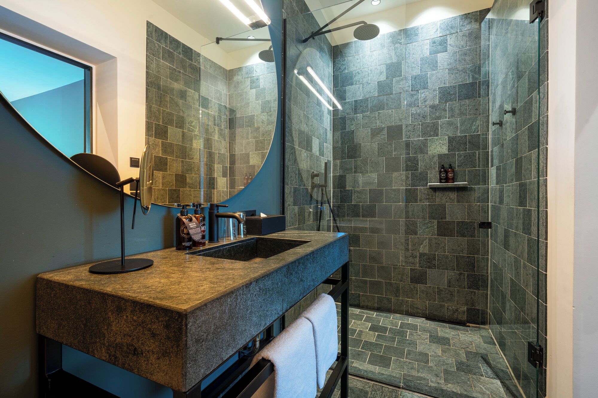 Un baño moderno con encimera de granito, un espejo grande y una zona de ducha con azulejos de piedra.