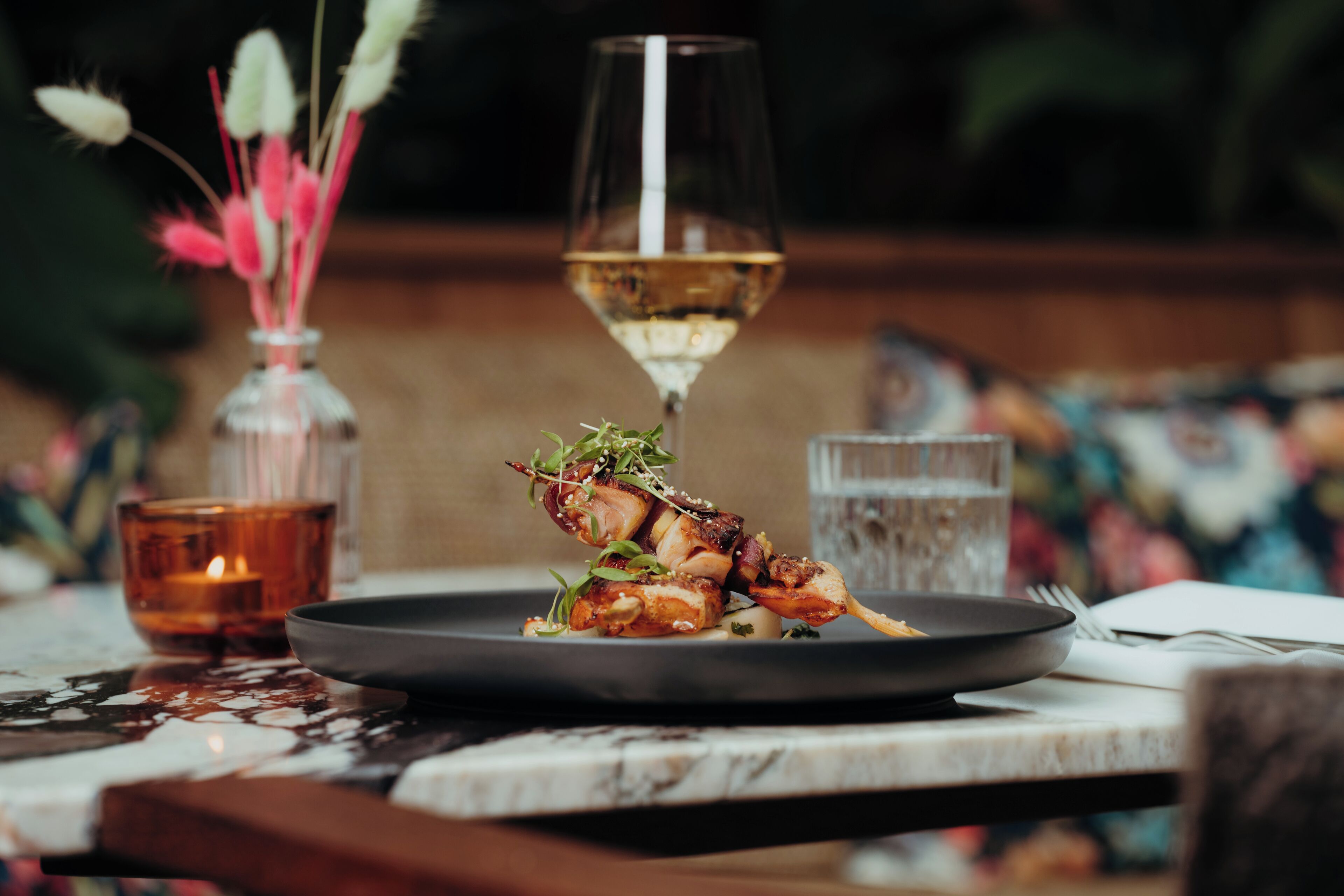 Une table à manger avec une assiette de nourriture, un verre de vin blanc et une bougie.
