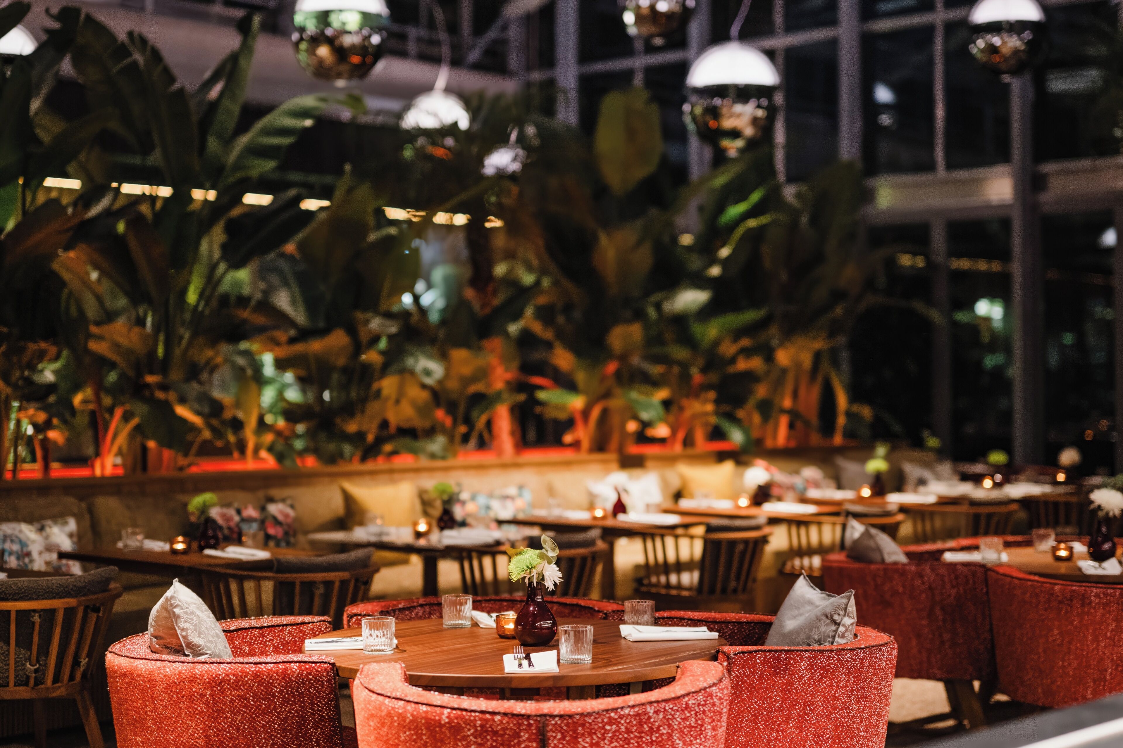 Restaurante com mesas redondas, cadeiras estofadas vermelhas e um painel de grandes plantas verdes.