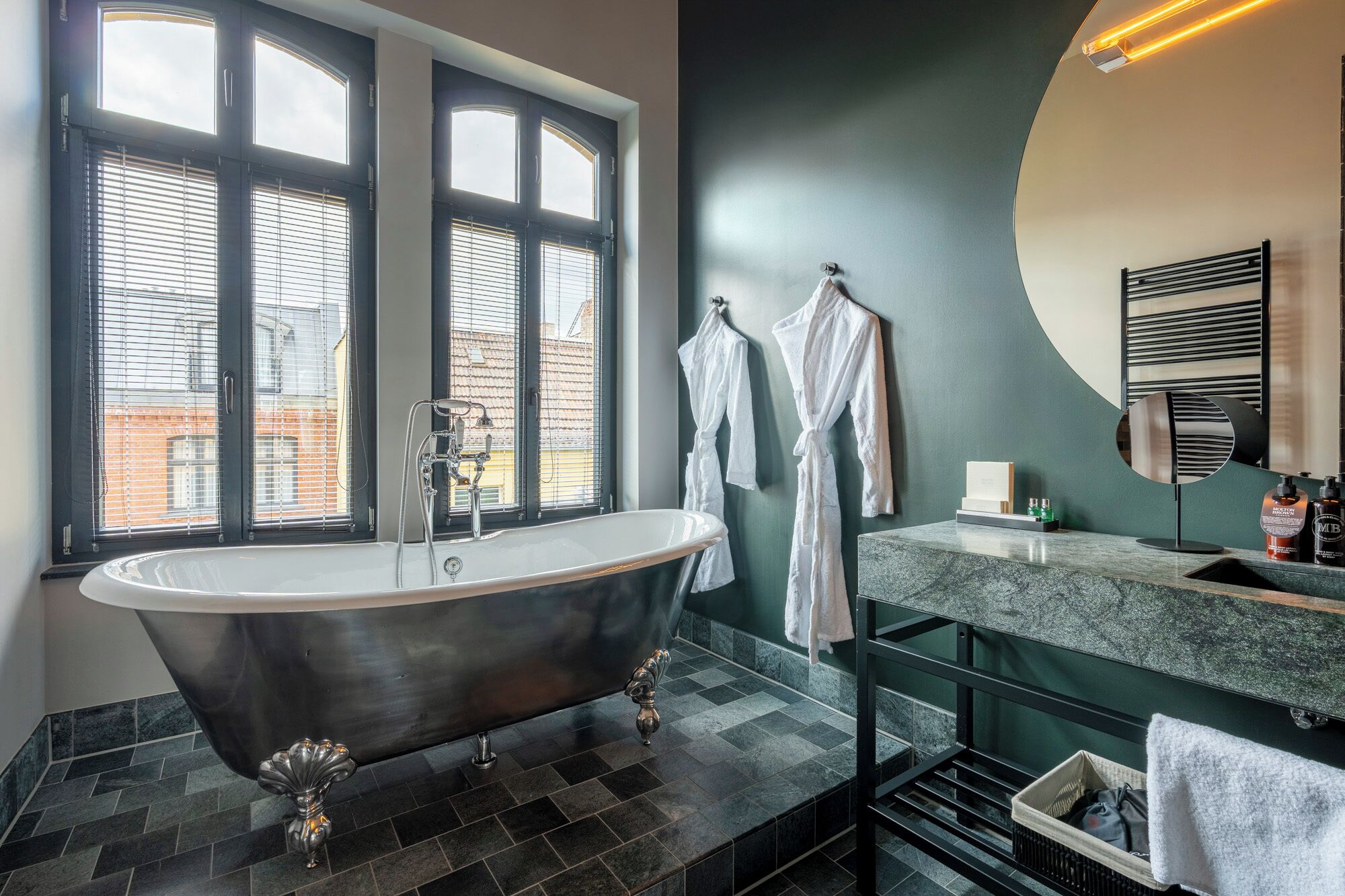 A modern bathroom with a freestanding bathtub, a large window with blinds, and a vanity with a mirror and toiletries.