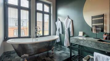 A modern bathroom with a freestanding bathtub, a large window with blinds, and a vanity with a mirror and toiletries.