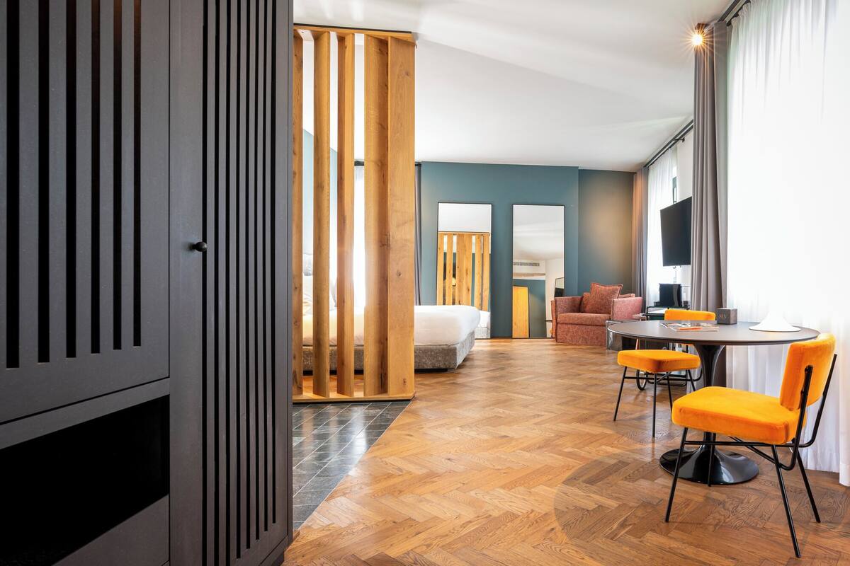 A modern hotel room with a wooden floor, a bed, a sofa, a dining table with chairs, and a large window with curtains.