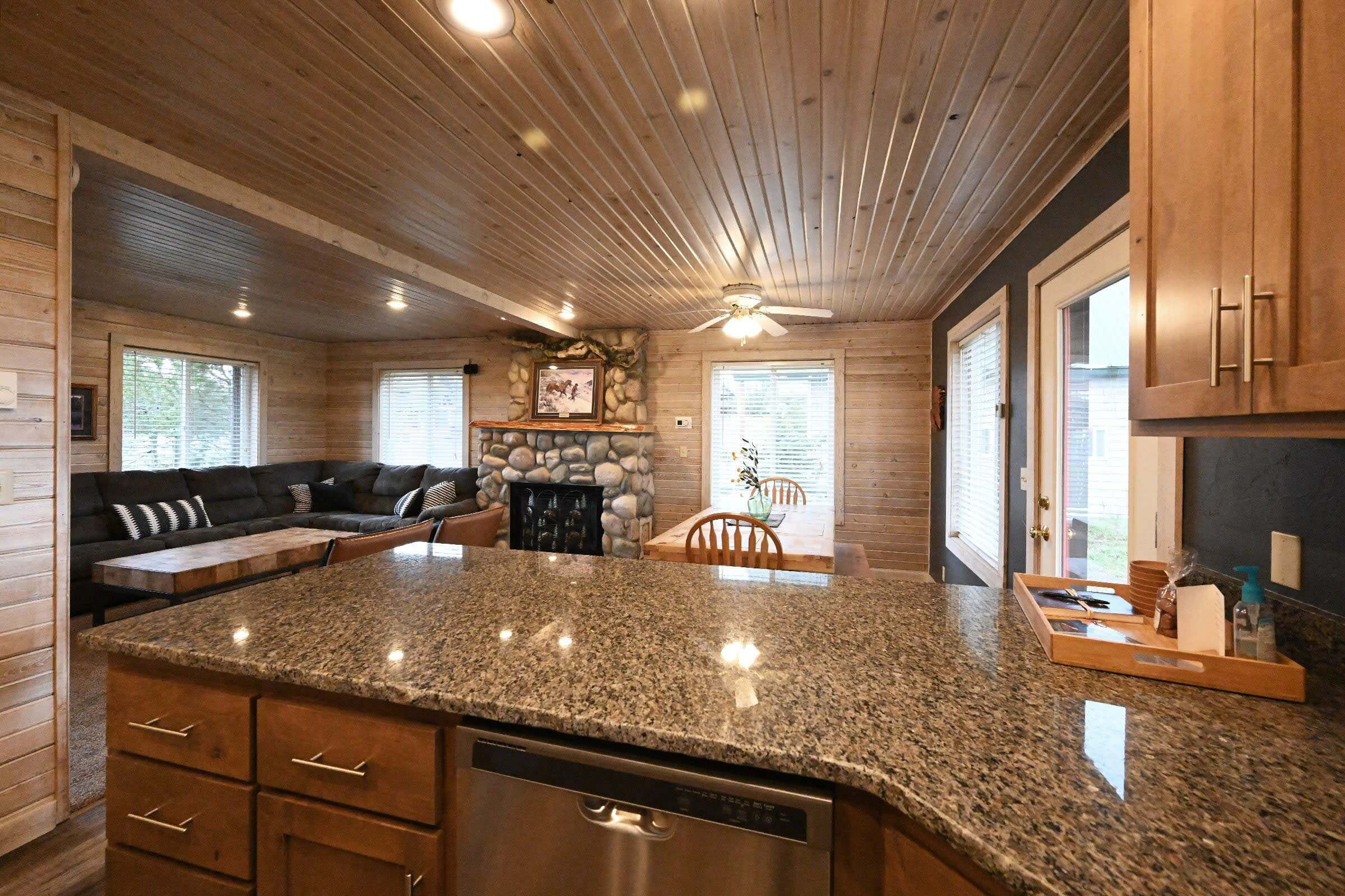 A kitchen with granite countertops, wooden cabinets, a built-in dishwasher, and a stone fireplace.
