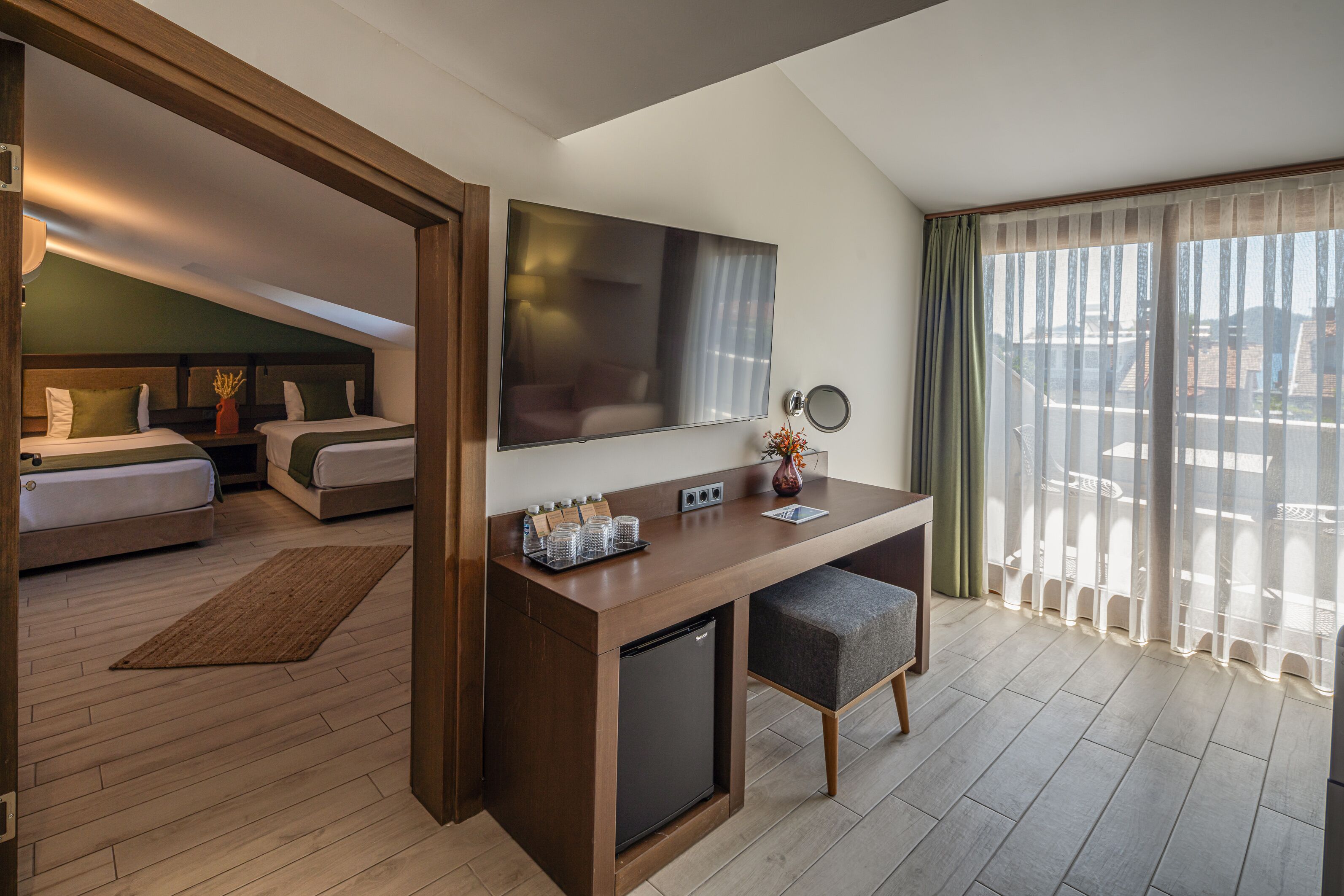 Une chambre d’hôtel équipée d’un bureau, d’un lit, d’une télévision, d’un petit réfrigérateur et d’un balcon avec des rideaux.